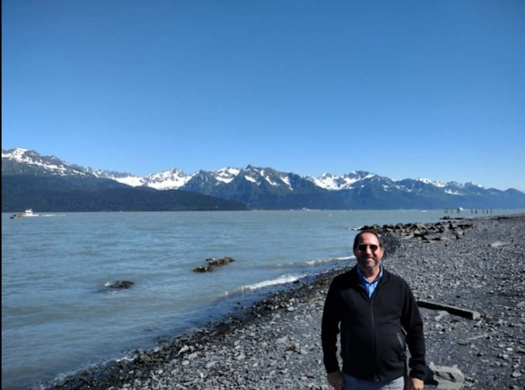 A person standing in front of a river with snow mountains in the far back