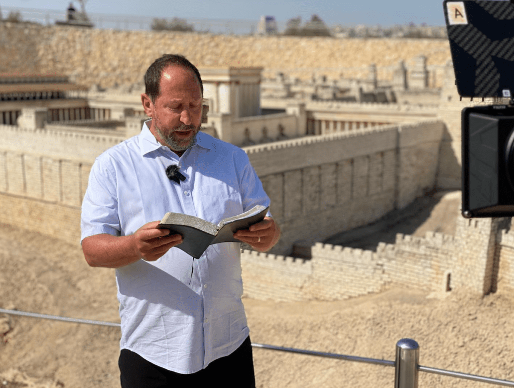 A person wearing a white shirt reading the bible in front of a structure.